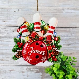 May include: A flat, hanging ornament featuring three white bowling pins with red stripes, a red bowling ball with the name James in white script, green pine branches, pinecones, red berries, and colorful string lights with a plaid bow.