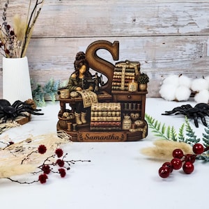 May include: A brown resin figurine featuring a large letter S, a person sewing at a desk, and a stack of books with the text 'just a girl who loves sewing' and 'Samantha' on the base. The scene includes miniature sewing supplies and a candle.