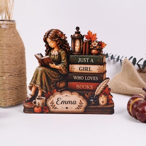 Può includere: Una statuetta decorativa che raffigura una persona intenta a leggere seduta su una pila di libri con la scritta Just a Girl Who Loves Books. La base include una targhetta con il nome Emma, una lanterna, una penna, una candela e foglie autunnali.