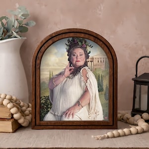 May include: A framed portrait of a woman in a white toga and floral crown. The portrait is set in a dark brown, arched wooden frame. The background of the portrait depicts a classical landscape with a building and trees.