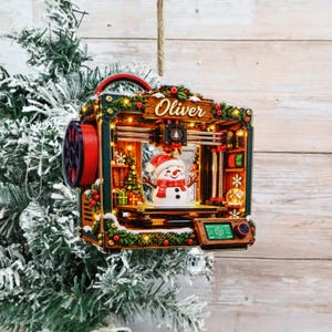 Pode incluir: Um enfeite de Natal em forma de impressora 3D com o nome Oliver, um boneco de neve central, guirlanda verde, bagas vermelhas e iluminação interna. A estrutura inclui uma bobina de filamento vermelha lateral e um pequeno ecrã digital.