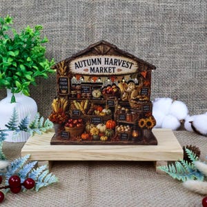 Puede incluir: Una miniatura de madera que representa un mercado de cosecha de otoño con estantes llenos de calabazas, manzanas y productos horneados. La estructura tiene un techo a dos aguas, luces y letreros con textos como Farm Fresh y Local Honey.