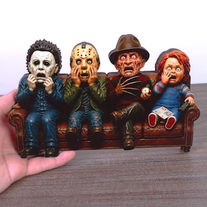 Op de afbeelding: Een decoratief beeldje met vier horrorfilmfiguren die op een bruine leren bank zitten. De personages zijn Michael Myers, Jason Voorhees, Freddy Krueger en Chucky, allemaal met geschokte gezichtsuitdrukkingen en hun handen voor hun gezicht.