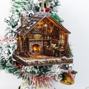 Puede incluir: Un adorno navideño colgante con forma de interior de cabaña con chimenea de piedra, sillón a cuadros y cabeza de ciervo. El letrero dice Blake's Hunting Lodge 2026. La escena presenta una iluminación interior cálida y una alfombra.