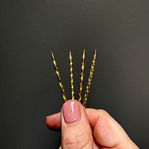 May include: Four gold-colored cocktail stirrers with a twisted design. The stirrers are held against a dark gray background. The tips of the stirrers are pointed. The image is a close-up shot.