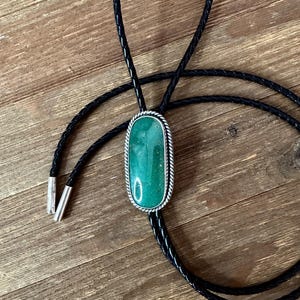 May include: A bolo tie featuring a rectangular, vibrant green stone set in a silver-toned frame with a braided rope design. The black leather cord is secured with silver-colored tips, creating a classic Western accessory.