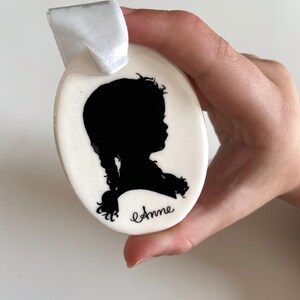 May include: White oval ceramic ornament with a black silhouette of a girl with a braid and the name "Anne" written in cursive.