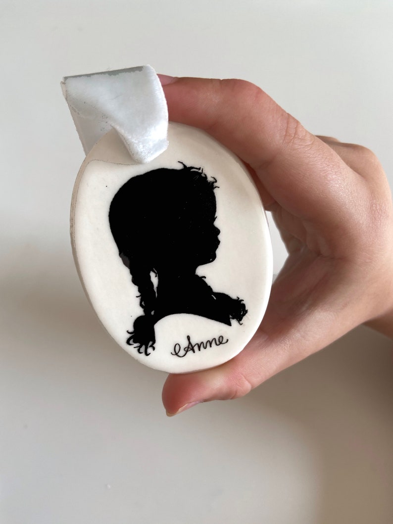 May include: White oval ceramic ornament with a black silhouette of a girl with a braid and the name "Anne" written in cursive.