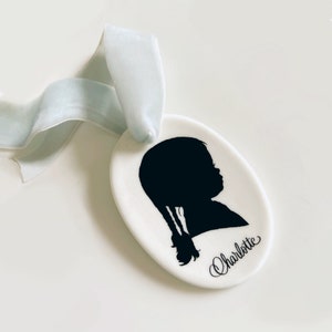 May include: White ceramic ornament with a black silhouette of a child with pigtails. The ornament has a blue ribbon and the name "Charlotte" written in cursive.