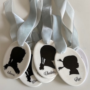 May include: Five white ceramic ornaments with black silhouette portraits of children. Each ornament has a blue ribbon attached and the name of the child is written below the portrait. The names are Anne, Don, Charlotte, and Pippi.