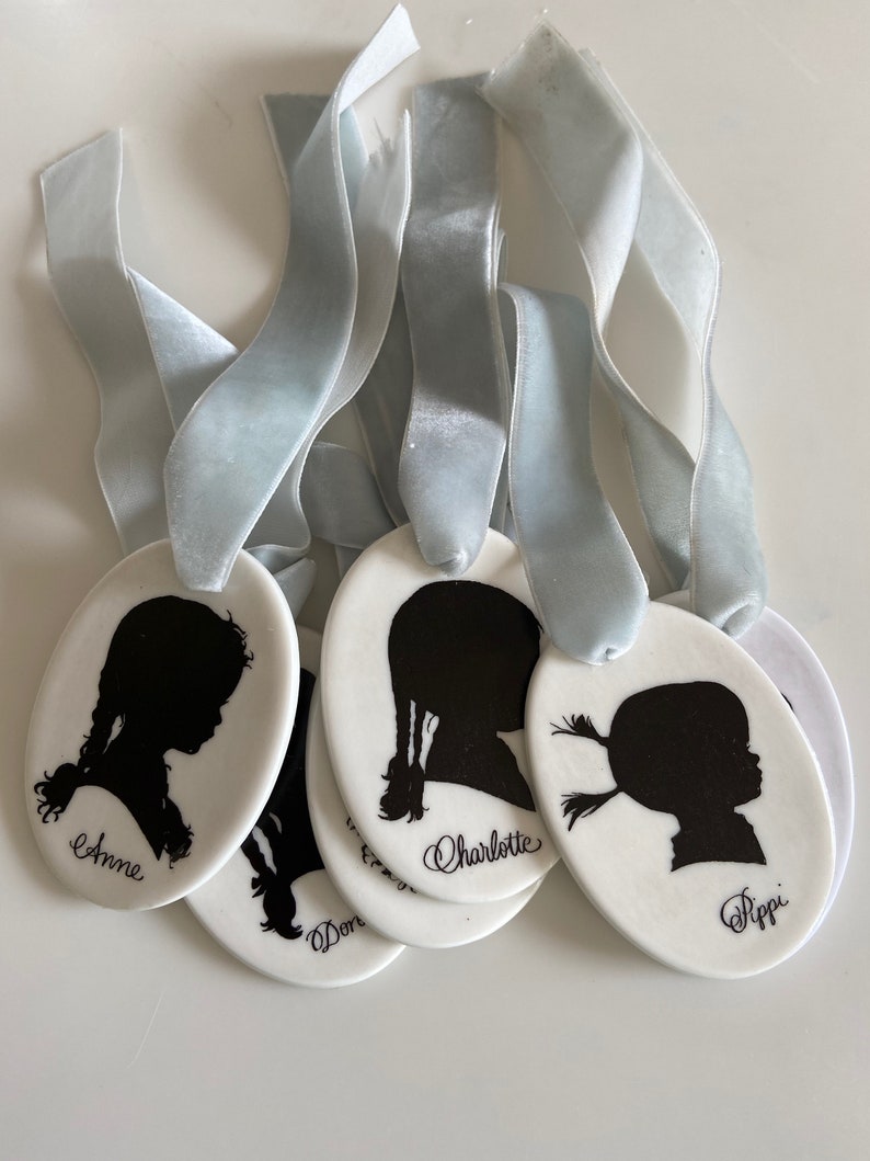 May include: Five white ceramic ornaments with black silhouette portraits of children. Each ornament has a blue ribbon attached and the name of the child is written below the portrait. The names are Anne, Don, Charlotte, and Pippi.