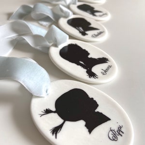 May include: Five white oval ceramic ornaments with black silhouettes of children's profiles and their names written in cursive. The ornaments are tied with light blue ribbon.