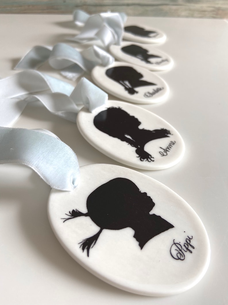 May include: Five white oval ceramic ornaments with black silhouettes of children's profiles and their names written in cursive. The ornaments are tied with light blue ribbon.