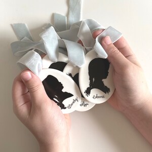 May include: Four white ceramic ornaments with black silhouette portraits and names "Anne", "Fippi", "Dorothy" and an unreadable name. The ornaments are held in a person's hands with light blue velvet ribbons.