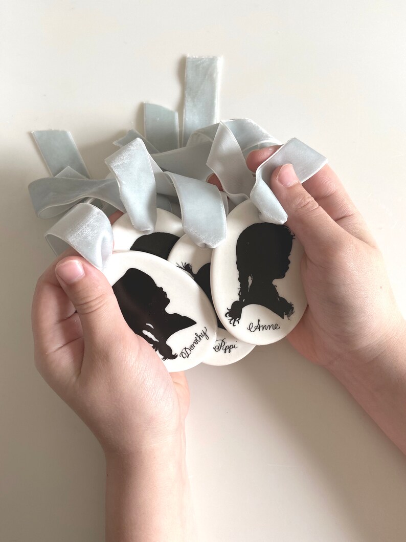 May include: Four white ceramic ornaments with black silhouette portraits and names "Anne", "Fippi", "Dorothy" and an unreadable name. The ornaments are held in a person's hands with light blue velvet ribbons.