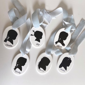 May include: Six white oval ceramic ornaments with blue ribbon hangers. Each ornament features a black silhouette of a girl's profile with a name written below: Charlotte, Pippi, Anne, Dorothy, Hermione, and Matilda.