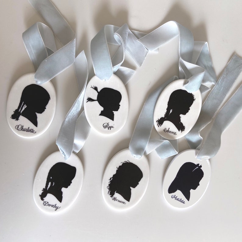 May include: Six white oval ceramic ornaments with blue ribbon hangers. Each ornament features a black silhouette of a girl's profile with a name written below: Charlotte, Pippi, Anne, Dorothy, Hermione, and Matilda.