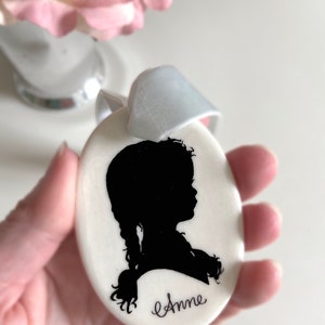 May include: White oval ceramic ornament with a black silhouette of a girl with a braid and the name "Anne" written in cursive.