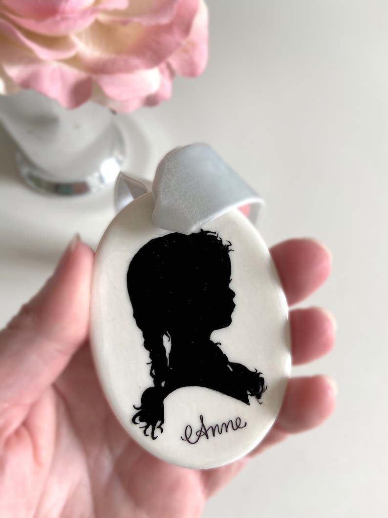 May include: White oval ceramic ornament with a black silhouette of a girl with a braid and the name "Anne" written in cursive.