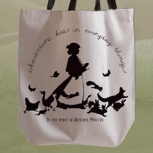 May include: A white tote bag with black handles featuring a silhouette design. The design includes a woman, animals, and the text "Adventure lies in everyday things." The phrase "In the spirit of Beatrix Potter" is also printed on the bag.