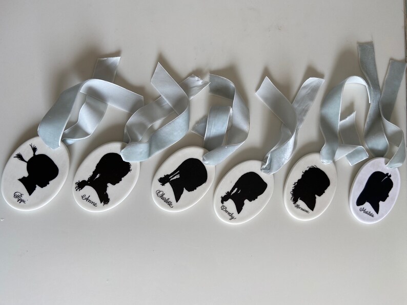May include: Seven white oval ornaments with blue ribbon ties. Each ornament features a black silhouette of a person's head and the name "Pippa", "Anne", "Charlotte", "Dorothy", "Hermione", "Amelia", and "Alisha" written in black.