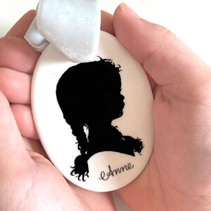 May include: White ceramic oval ornament with a black silhouette of a girl with a braid and the name "Anne" written in cursive.