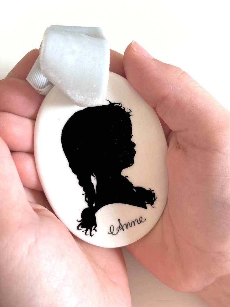 May include: White ceramic oval ornament with a black silhouette of a girl with a braid and the name "Anne" written in cursive.