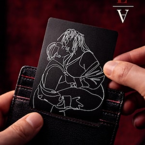 May include: A black card with a white line drawing of two people kissing, held in a black wallet. The card has the letters "L" and "V" with a lipstick mark above the "L".