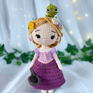 Puede incluir: Una muñeca de crochet con el pelo largo y rubio, que lleva un vestido morado y una corona de flores. Un pequeño camaleón de crochet verde se sienta en la cabeza de la muñeca. La muñeca sostiene una pequeña sartén de crochet gris.