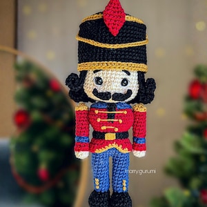 Könnte beinhalten: Handgefertigte gehäkelte Nussknacker-Puppe. Die Puppe hat einen schwarzen Hut mit goldenem Besatz und einem roten Diamant-Akzent. Die Jacke ist rot mit goldenem Besatz und die Hose ist blau. Die Puppe hat ein weißes Gesicht mit einem schwarzen Schnurrbart und schwarzen Locken.