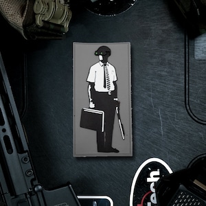 Może przedstawiać: Czarno-biała naszywka przedstawiająca osobę w hełmie i okularach, trzymającą teczkę i laskę. Naszywka znajduje się na szarym tle.