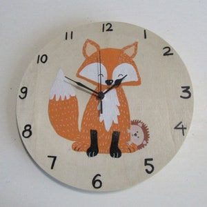 May include: A round wooden clock featuring a cartoon fox with orange fur, white accents, and black feet. The clock face has black numerals and hands. A small hedgehog is also visible. The clock is designed for a nursery or children's room.