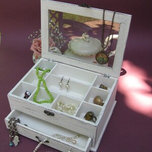 May include: White wooden jewelry box with a mirrored lid and a drawer. The box has multiple compartments for storing jewelry, including rings, earrings, necklaces, and bracelets.