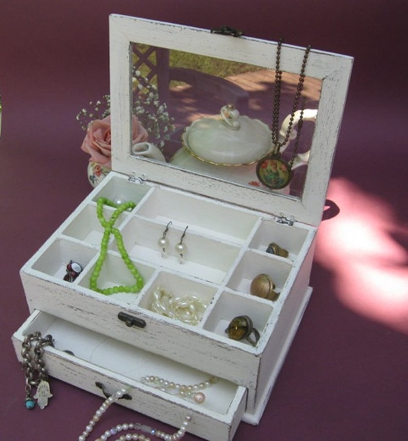 May include: White wooden jewelry box with a mirrored lid and a drawer. The box has multiple compartments for storing jewelry, including rings, earrings, necklaces, and bracelets.