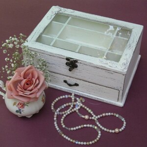 May include: White wooden jewelry box with a glass top and a drawer. The box has a distressed finish and is decorated with floral carvings. A pink rose is in a small porcelain pitcher next to the box, and a pearl necklace is on the table.