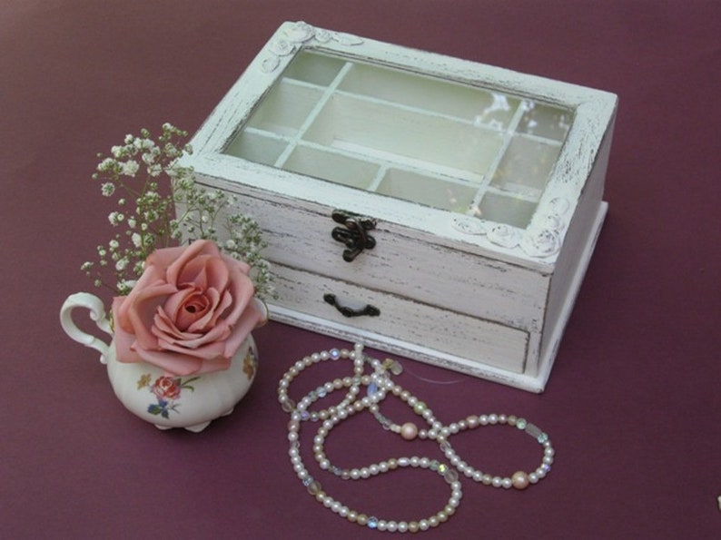 May include: White wooden jewelry box with a glass top and a drawer. The box has a distressed finish and is decorated with floral carvings. A pink rose is in a small porcelain pitcher next to the box, and a pearl necklace is on the table.