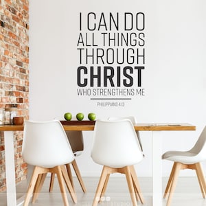 Op de afbeelding: Een witte muur met een zwarte vinyl sticker waarop staat "Ik kan alles door Christus, die mij kracht geeft, doen Filippenzen 4:13". De sticker bevindt zich boven een houten tafel met vier witte stoelen.