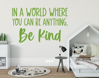 May include: Green wall decal with the text "IN A WORLD WHERE YOU CAN BE ANYTHING, Be Kind" in a white room with a white bed and a white nightstand.