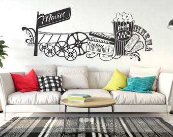 May include: Black and white movie themed wall decal with a sign that says "Movies", film reels, a clapperboard, popcorn, and tickets. The decal is on a white wall above a white couch with colorful pillows.
