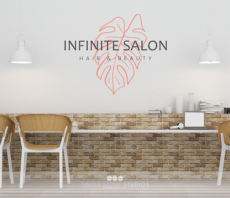 Pu&ograve; includere: Un muro bianco con un muro di mattoni dietro un bancone bianco. Il muro presenta un disegno di foglia rosa con il testo "INFINITE SALON HAIR & BEAUTY" e il testo "SINGLE STONE STUDIOS DESIGN & BUILD" in basso.