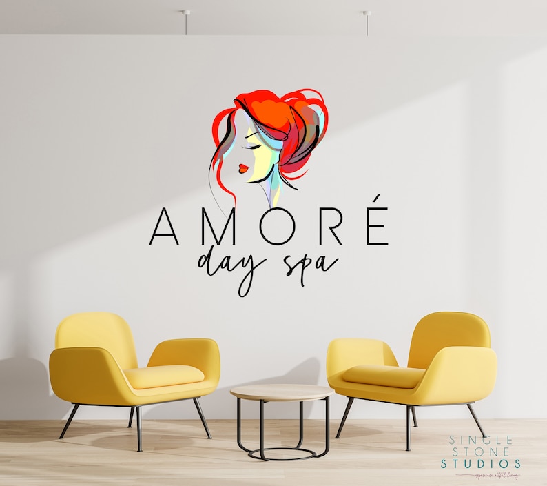 Pu&ograve; includere: Un muro bianco con un logo per una spa diurna chiamata "AMOR&Eacute; day spa". Il logo presenta un'illustrazione colorata del viso di una donna. Due poltrone gialle e un piccolo tavolo di legno si trovano di fronte al muro.