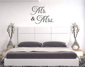 May include: A white headboard with a black vinyl decal that reads "Mr. & Mrs.". The headboard is on a bed with gray bedding and a white frame. The bed is in a bedroom with a white wall and a gray rug.