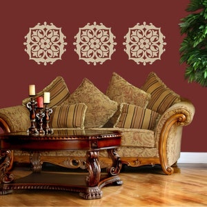 Op de afbeelding: Een bruine, sierlijke bank met drie kussens staat voor een bordeauxrode muur met drie decoratieve stencils. De stencils zijn beige en hebben een bloemmotief. Een kleine, ronde, houten salontafel staat voor de bank. Een plant in een pot staat in de hoek van de kamer.