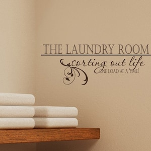Op de afbeelding: Een bruine muur met een wit rek en een stapel witte handdoeken. De muur heeft een bruine vinyl sticker met de tekst "The Laundry Room sorting out life one load at a time!"