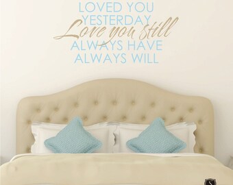 May include: A wall decal with the text "Loved you yesterday, Love you still, Always have, Always will" in blue and brown lettering. The decal is displayed above a beige headboard with two light blue patterned pillows.