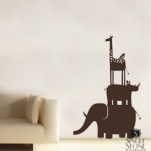 May include: A brown silhouette wall decal of an elephant, rhinoceros, and giraffe stacked on top of each other. The giraffe has a small bird perched on its back. The decal is on a white wall with a white couch in the foreground.