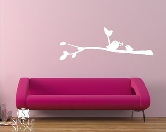 May include: White vinyl wall decal of two birds perched on a branch with leaves. The decal is on a pink wall above a pink couch.
