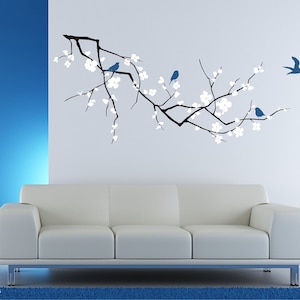 Op de afbeelding: Een witte bank staat voor een muur met een zwart-wit silhouet van een kersenbloesemtak met blauwe vogels. De muur is aan de linkerkant blauw en aan de rechterkant wit geverfd.