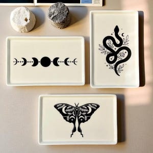 Puede incluir: Juego de tres bandejas rectangulares color crema con diseños gráficos negros. Una bandeja presenta una serpiente con elementos florales, otra una polilla y la tercera un diseño de fases lunares. Ideal para servir o exhibir.