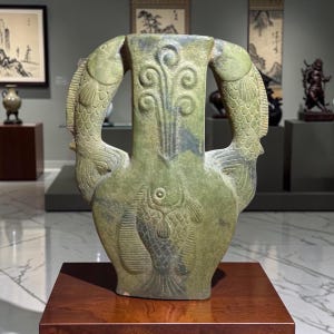 May include: A light green ceramic vase featuring a central fish relief on the body and two large fish forming the handles on either side. The neck of the vase is decorated with a vertical, symmetrical scroll pattern carved into the surface.